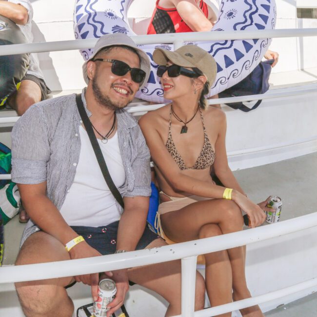 A man and woman sit close together on a boat, smiling and wearing sunglasses. The man holds a drink and wears a hat and casual clothes; the woman wears a bikini and a cap. Inner tubes and other people are in the background.