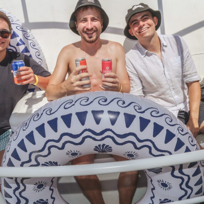 Three smiling men sit behind a large inflatable pool float on a boat. Two hold cans of soda or beer, and all wear summer clothing and sunglasses. Sunlight and a relaxed, festive atmosphere are evident.