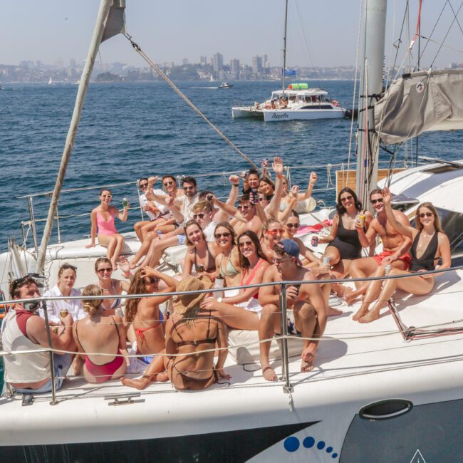 A group of young adults in swimsuits relax and pose for a photo on the deck of a sailboat, smiling and waving, with the ocean and city skyline visible in the background on a sunny day.