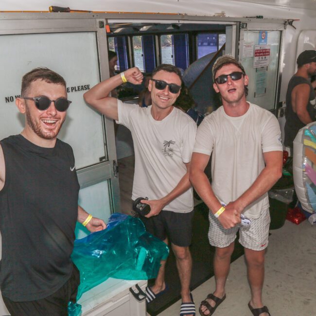 Three men in casual summer clothes and sunglasses flex and pose for the camera on a boat party. Other people are in the background, some holding drinks and wearing swimsuits. The scene is lively and festive.