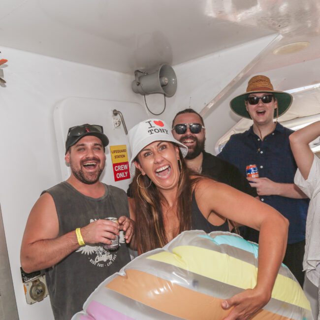 A group of five adults laughing and smiling on a boat. One woman holds a colorful pool float and wears an “I ❤️ TONY” hat. Others wear sunglasses, summer clothes, and hats, enjoying a lively atmosphere.