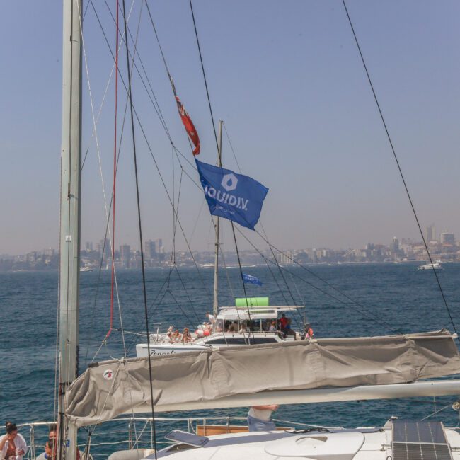 A group of people on a boat with a blue "LIQUIDIX" flag, sailing on a body of water with a city skyline visible in the background under a hazy sky.