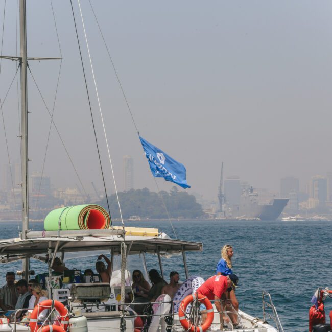 A group of people relax on a sailing yacht with a blue flag on open water, while someone takes a photo. The coastline and city skyline are visible in the hazy background.