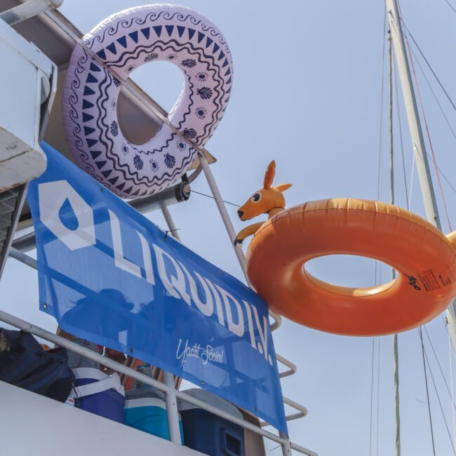 A yacht with a blue "LIQUIDITY" banner displays two inflatable swim rings, one patterned and one orange with a kangaroo head, hanging from the upper deck against a clear sky.