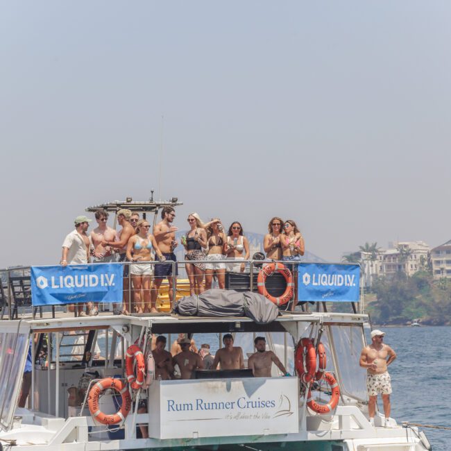 A group of people in swimsuits stand and socialize on the upper deck of a boat labeled "Rum Runner Cruises," with banners for "LIQUID I.V." The boat is on the water, with buildings and trees visible in the background.