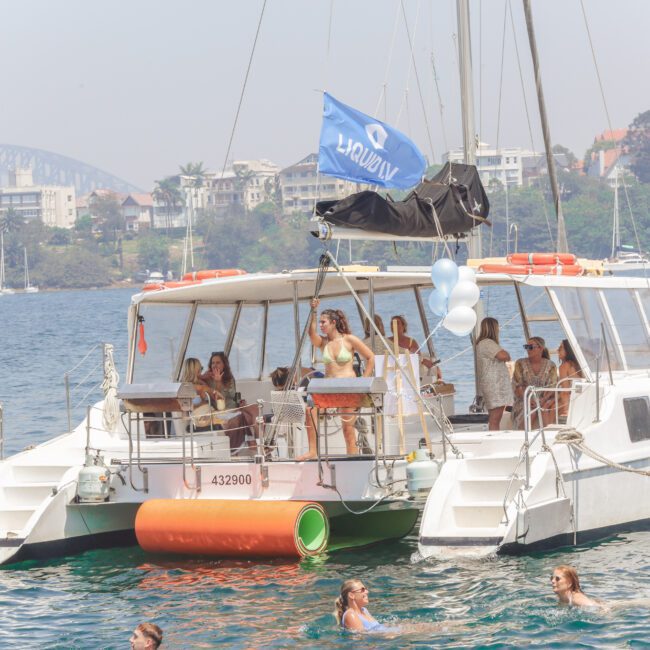 A group of people are gathered on a white catamaran in a harbor, some standing and some sitting, with a blue flag, balloons, and a cityscape with trees and buildings visible in the background.