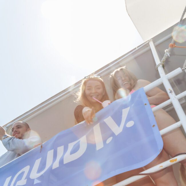 Three people stand on a boat deck, smiling and looking down over a railing draped with a blue banner. One person is taking a photo with a phone. The sun shines brightly, creating lens flares in the image.
