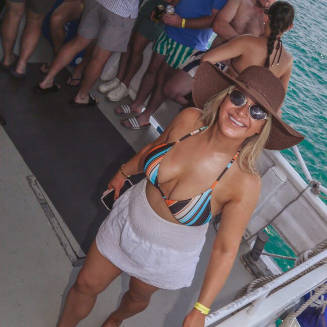 A smiling woman in a colorful bikini top, white skirt, sunhat, and sunglasses stands on a boat deck near a railing. Other people are socializing in the background. Blue-green water is visible beside the boat.