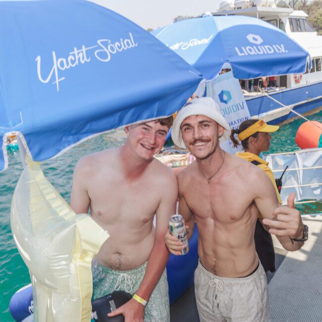 Two smiling shirtless men pose together on a yacht party, holding drinks. Other people, umbrellas, and a large boat are visible in the background on bright blue water.