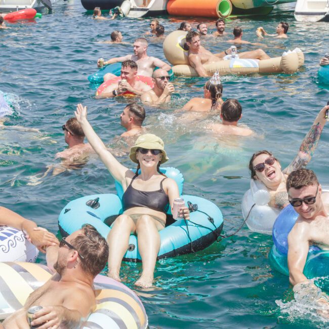 A group of people relax on colorful inflatables, smiling and holding drinks, while floating together in clear blue water near boats on a sunny day.