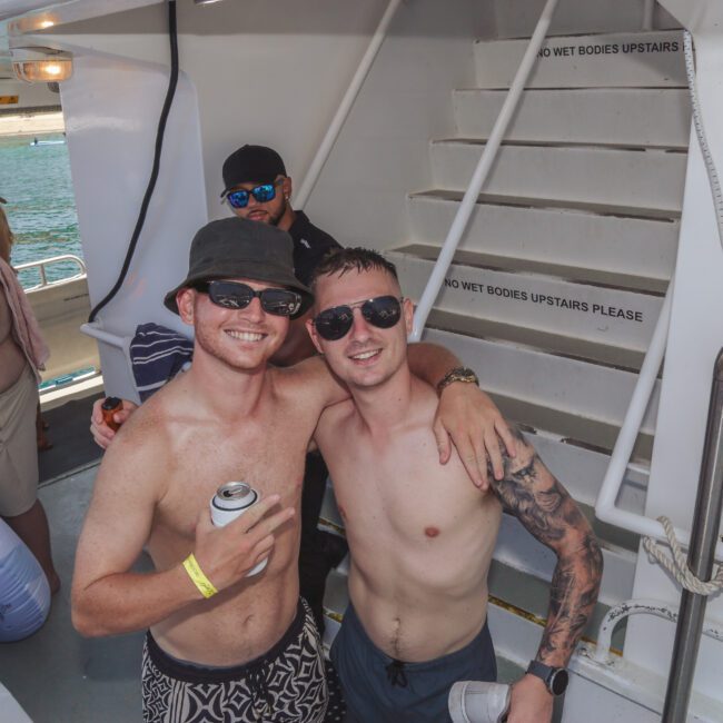 Two smiling men in swimsuits and sunglasses pose arm-in-arm on a boat. Other people relax nearby, some holding drinks or wrapped in towels. The scene is sunny and festive, with water and coastline visible in the background.
