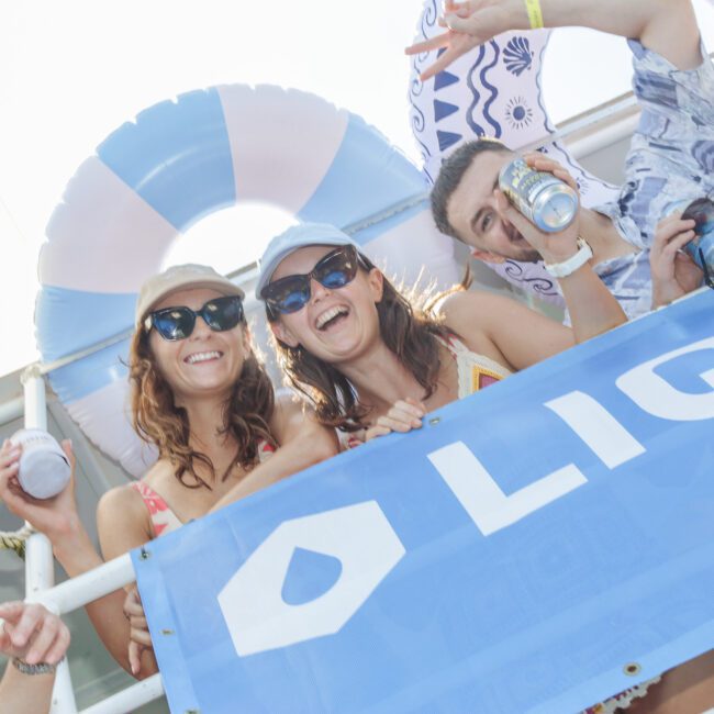 Three people in sunglasses and hats smile and hold drinks on a boat, standing behind a blue banner. A large inflatable ring is in the background, and it’s a sunny day.