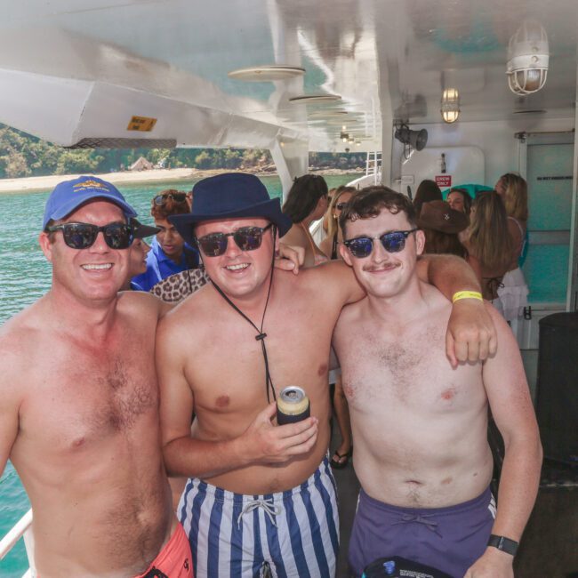 Three smiling men wearing sunglasses and swim trunks pose together on a boat. The ocean and green hills are visible in the background, and other people can be seen enjoying the sunny day onboard.