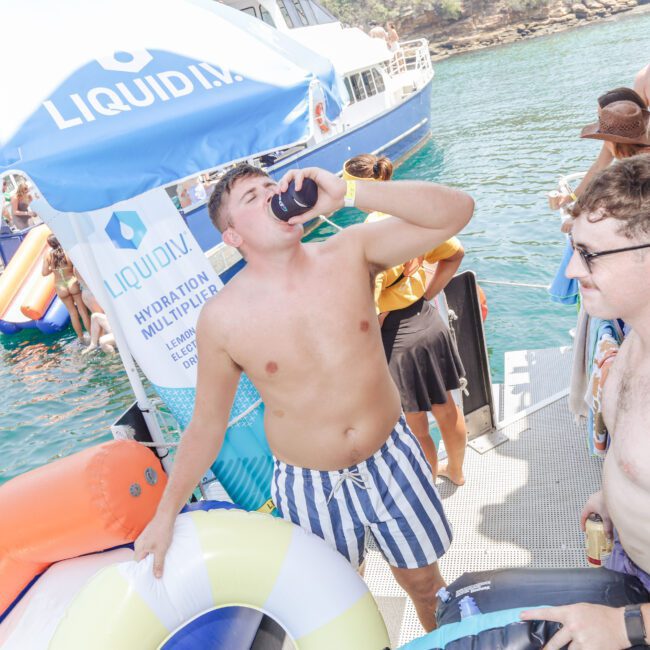 A group of people in swimsuits enjoy a sunny day on a boat and dock. One man in striped shorts drinks from a can, while others stand nearby. Inflatable pool floats and a boat with a "LIQUIDN" banner are visible.