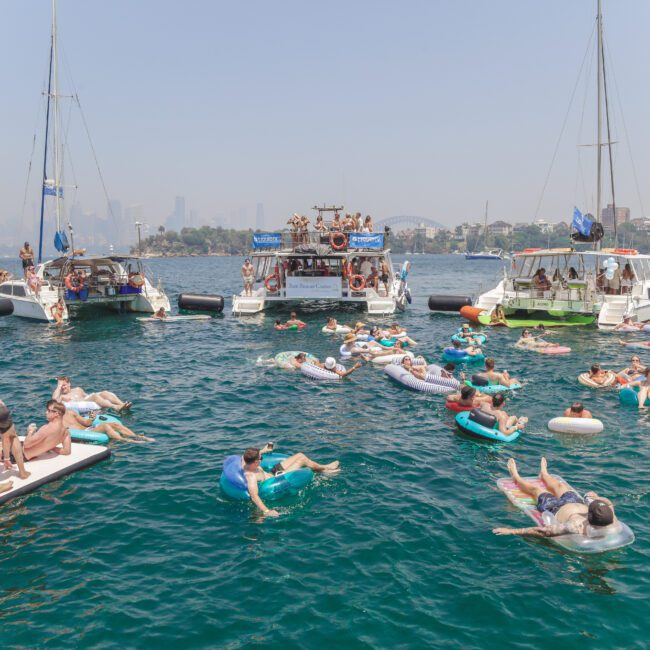 People relax on inflatable tubes and mats in the water near anchored boats during a sunny day party. The background shows a city skyline and the Sydney Harbour Bridge under a hazy sky.