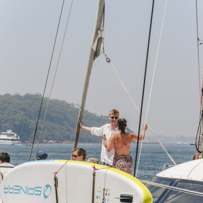 Two people stand and talk on the deck of a sailboat, with one man in a white shirt and sunglasses smiling. A paddleboard is attached to the boat, and the sea and distant hills are visible in the background.