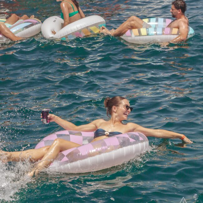 A woman in sunglasses lounges on a pastel inflatable ring, smiling and holding a drink, while others float nearby in clear blue water on a sunny day. The logo "Yacht Social Club" is visible in the corner.