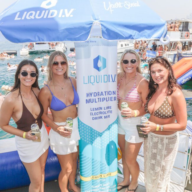 Four women in swimsuits and skirts stand smiling on a boat next to a large Liquid I.V. display. Boats and people swimming are visible in the background on a sunny day. The scene appears festive and summery.