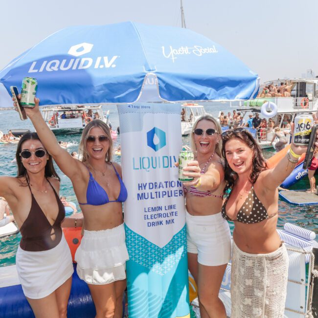 Four women in swimsuits and sunglasses smile and raise drinks by a blue Liquid I.V. umbrella at a lively boat party on the water, with other people and boats in the background on a sunny day.