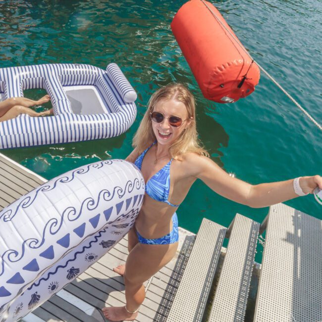 A smiling woman in a blue bikini and sunglasses stands on a dock by turquoise water, holding a white inflatable float. Other inflatables and a large orange buoy are nearby. She appears ready to enjoy a swim.