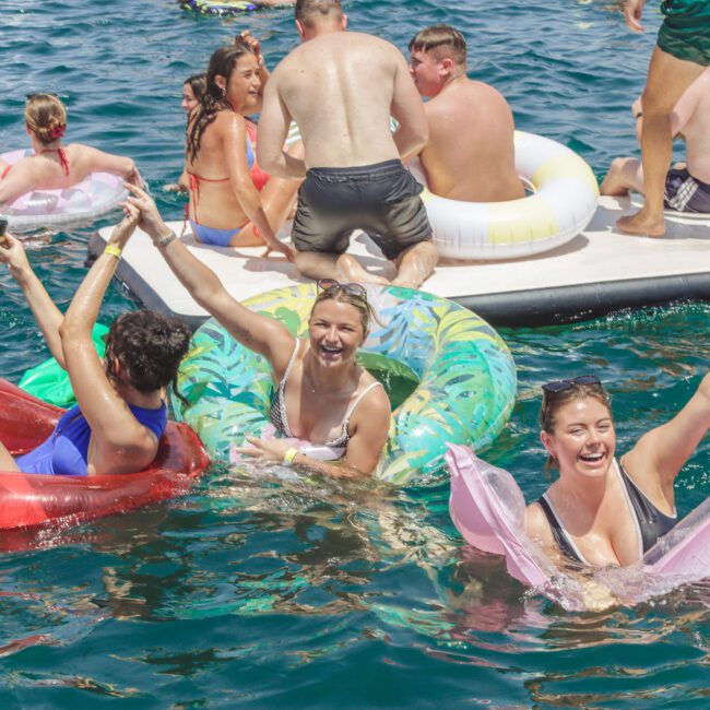 A group of people enjoy a sunny day on the water, floating on colorful inflatables. Some are posing and smiling for the camera, while others relax or talk on a floating dock in the background.