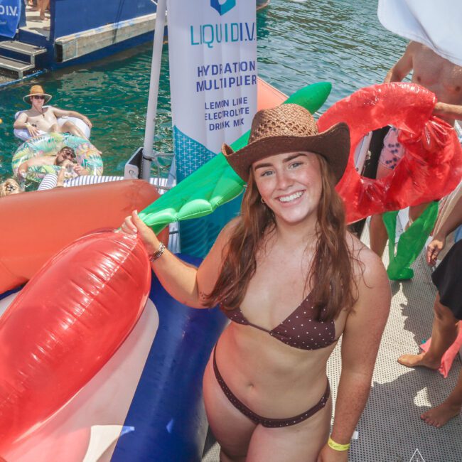 A smiling woman in a brown polka-dot bikini and cowboy hat stands on a dock near large inflatable pool toys, with water and people in the background. A hydration drink banner is visible behind her.