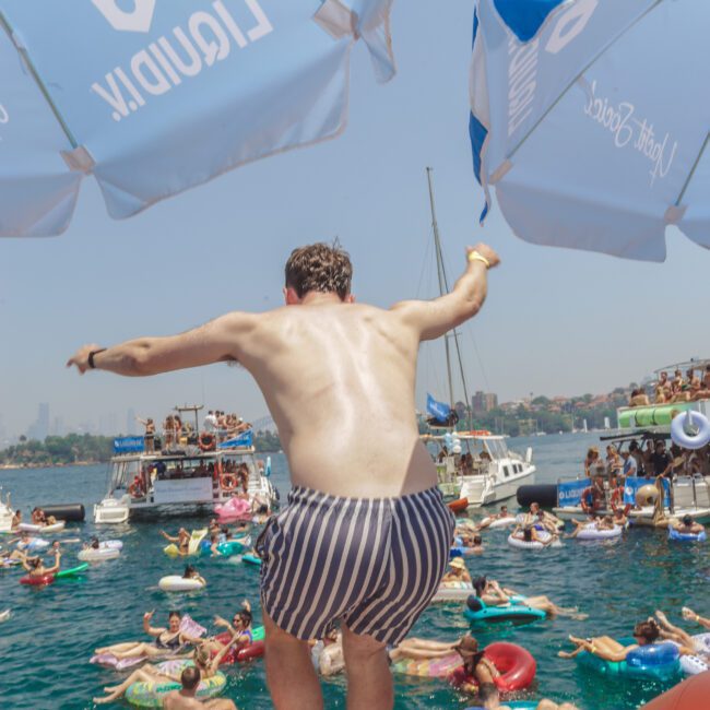 A man in striped swim shorts jumps from a floating platform into a crowded, vibrant water party with people on inflatables, surrounded by yachts and blue umbrellas under a sunny sky.
