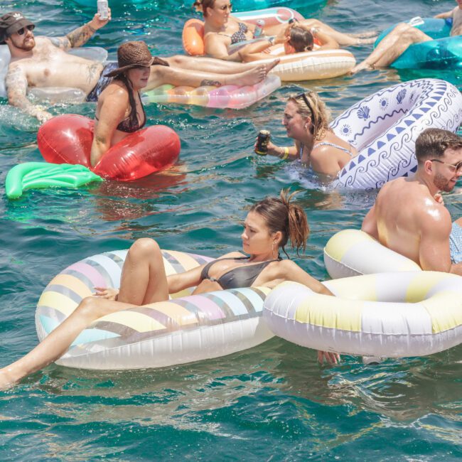 People relax on colorful inflatable pool floats in clear blue water, some holding drinks and chatting, enjoying a sunny day. Most wear swimsuits and sunglasses. The atmosphere appears lively and fun.