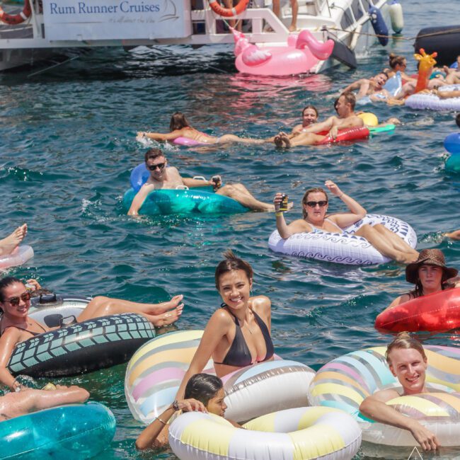 People relaxing on colorful inflatable tubes in the water near a boat labeled "Rum Runner Cruises," enjoying a sunny day. Some hold drinks, and everyone appears to be having fun.