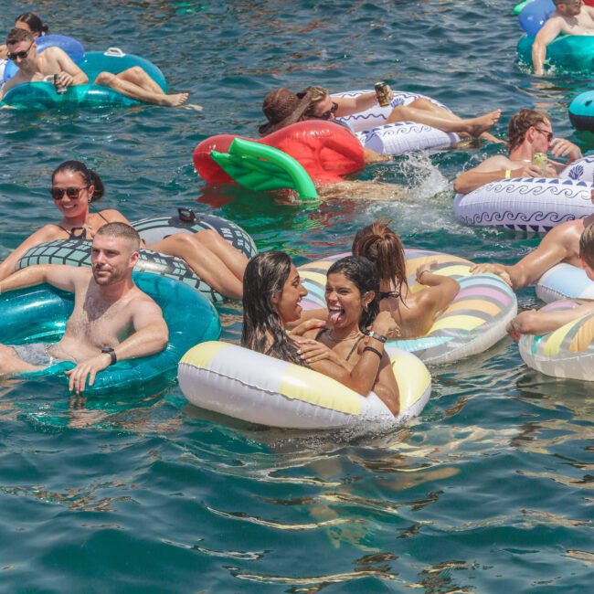 A group of people relax and have fun on colorful inflatable pool floats in the water during a sunny day, with a “Yacht Social” logo in the corner of the image.