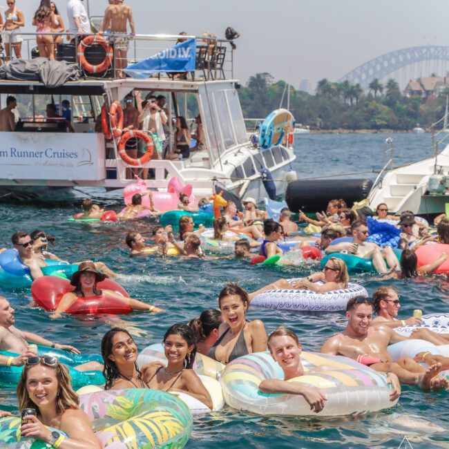A large group of people relax on colorful inflatable tubes in the water near anchored boats, with the Sydney Harbour Bridge visible in the background on a sunny day.