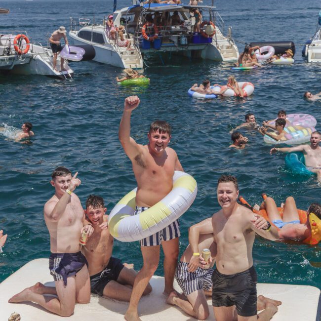 Four young men in swimsuits pose cheerfully on a floating mat in the water, surrounded by other people swimming, relaxing on inflatables, and boats. A city skyline is visible in the background through haze.