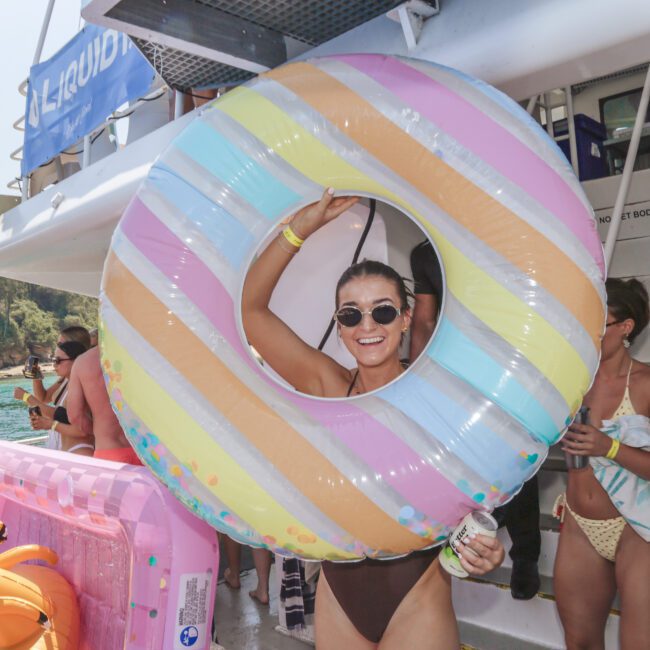 A smiling woman in sunglasses holds a large, colorful striped inflatable swim ring on a boat surrounded by people in swimwear, with water and trees visible in the background.