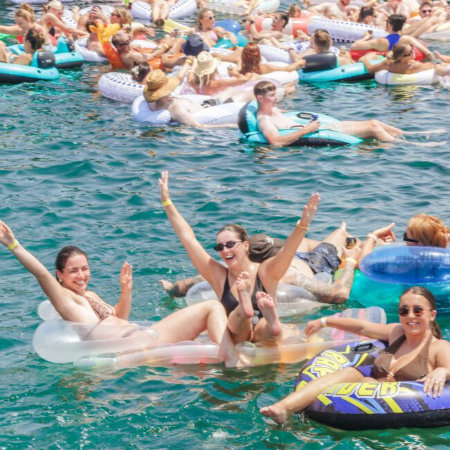 A large group of people float on colorful inflatable tubes in bright blue water, smiling and waving at the camera during a lively outdoor event under sunny skies.