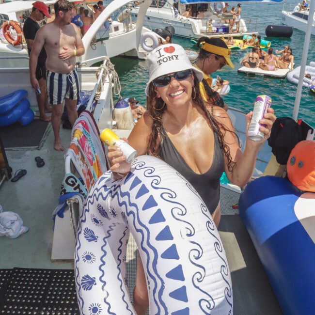 A woman in sunglasses and a white “I ❤️ TONY” hat smiles while holding drinks and a float, surrounded by people, inflatables, and boats on the water at a lively summer party.