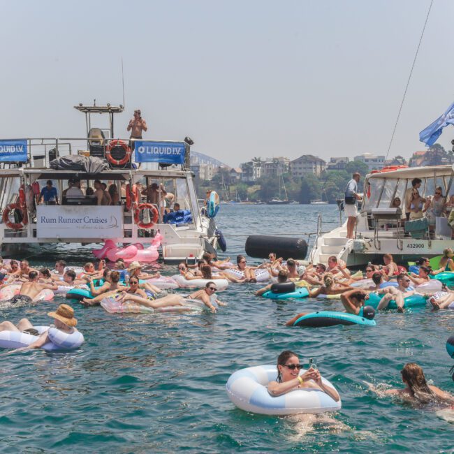 A large group of people relax on colorful inflatables in the water between two boats on a sunny day, with city buildings and trees visible in the background.
