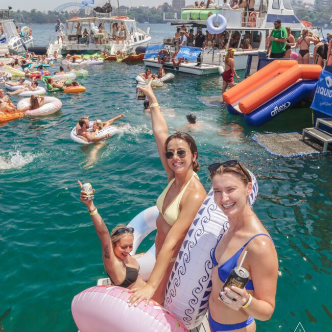 Three women in swimsuits smile and raise drinks on inflatable pool floats in the water near anchored party boats, surrounded by people swimming and sunbathing at a lively yacht party. Blue sky and city skyline in the background.