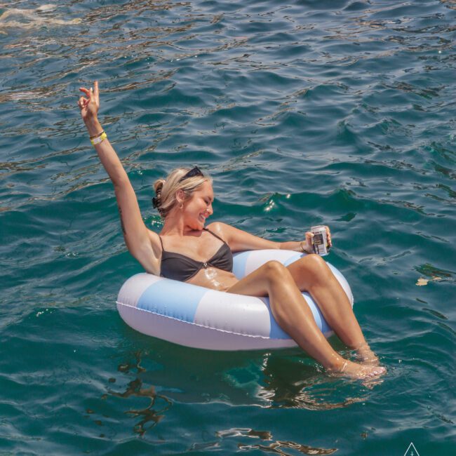 A woman in a black bikini relaxes on a blue and white inflatable ring in the water, holding a drink and raising one arm cheerfully. The sun reflects on the rippling water. "Yacht Social Club" logo is visible in the corner.