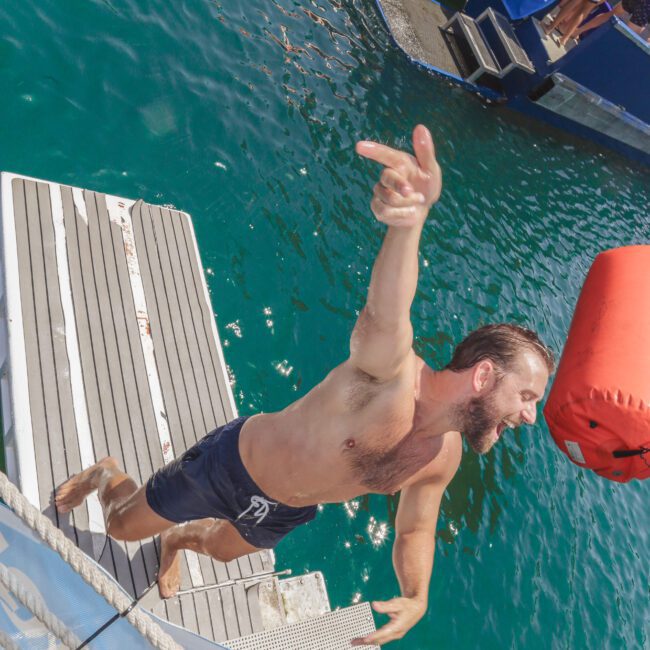 A man in swim trunks slips off a metal boat ladder, mid-fall into clear blue-green water, with one arm raised. Floating docks and other people are visible in the background. The scene is lively and sunlit.