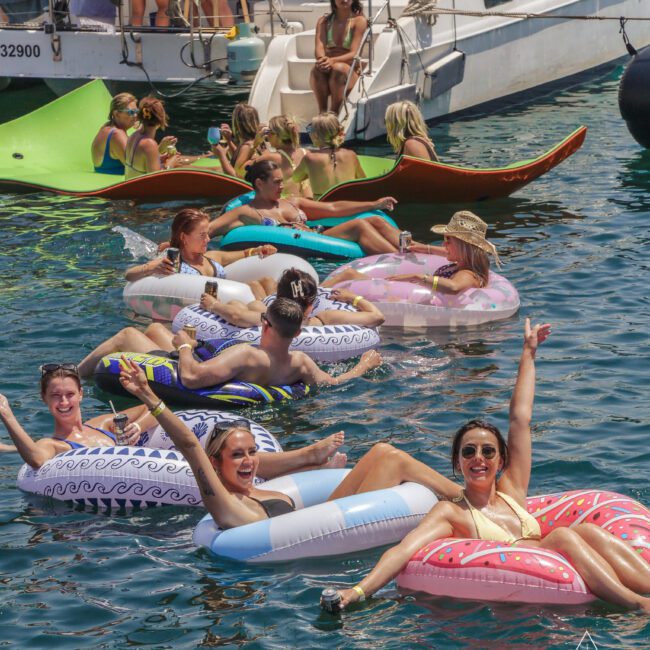 A group of people relax and smile on colorful inflatable floats in the water near a boat, enjoying a sunny day. Some wave at the camera while others lounge, with more people sitting on the yacht in the background.