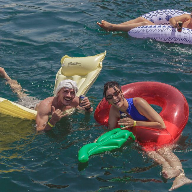 Two people smile and pose in the water, floating on inflatable pool toys—one red ring and one yellow mat—while holding drinks. Others on inflatables are visible in the background. Logo at the bottom reads “The Yacht Social Club.”.