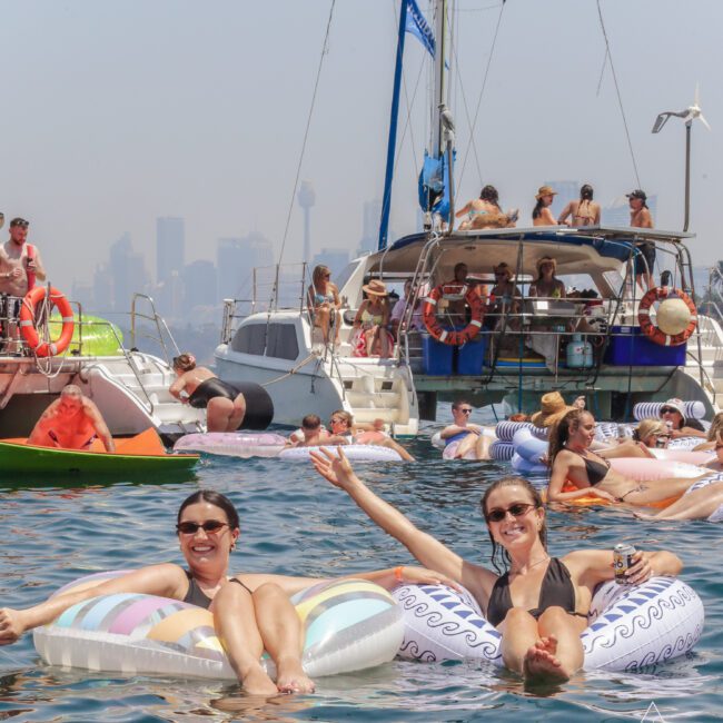 Two smiling women relax on colorful pool floats in the water, surrounded by boats and other people enjoying a sunny day. The city skyline is visible in the hazy background.