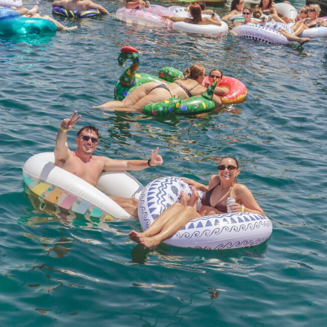 People relax on colorful inflatable floats in clear blue water near boats. A smiling man and woman in sunglasses pose and make peace signs in the foreground. Others lounge and socialize on floats and on a docked yacht in the background.