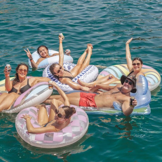 Six people in swimsuits relax on colorful pool floats in blue water, smiling and raising drinks. The group appears to be enjoying a sunny day, with the "Yacht Social Club" logo in the bottom right corner.