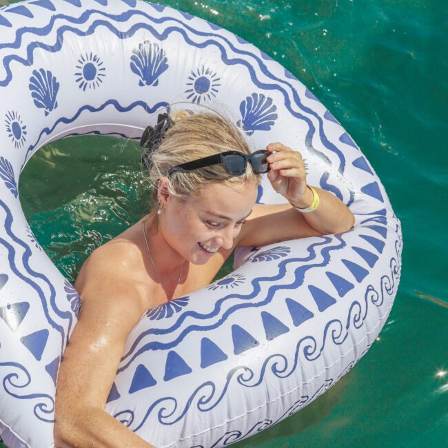 A woman with blonde hair relaxes in turquoise water on a decorative pool float, smiling as she holds a can and adjusts her sunglasses. The scene is bright and sunny, with splashes of water around her.
