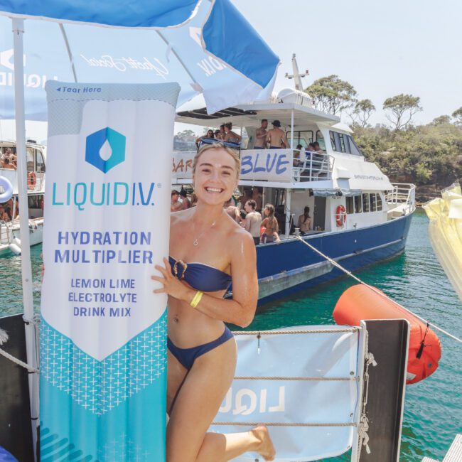 A smiling woman in a blue bikini stands on a dock, hugging a large inflatable Liquid I.V. Hydration Multiplier drink mix package. A boat with people on board and trees are visible in the background.