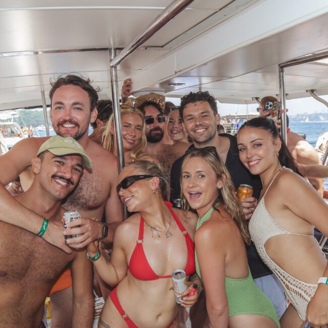 A group of smiling young adults in swimsuits pose closely together on a boat, enjoying drinks, with the sea and other boats visible in the background on a sunny day.