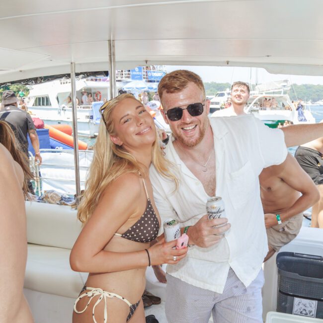 A smiling woman in a brown polka-dot bikini and a man in a white shirt with sunglasses hold drinks and pose together on a lively boat, surrounded by other people and boats in the background on a sunny day.