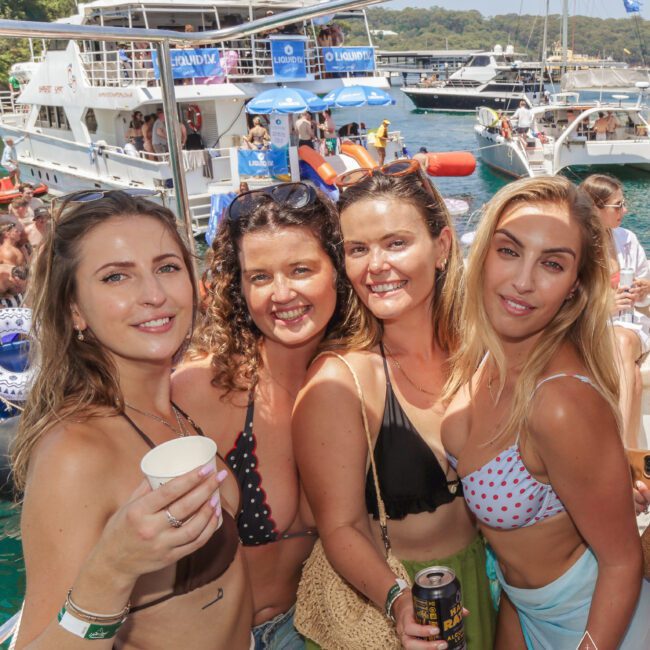 Four women in swimsuits smile and pose together at a lively boat party, with other people, boats, and water visible in the sunny background. One woman holds a drink and the Yacht Social Club logo is on the image.