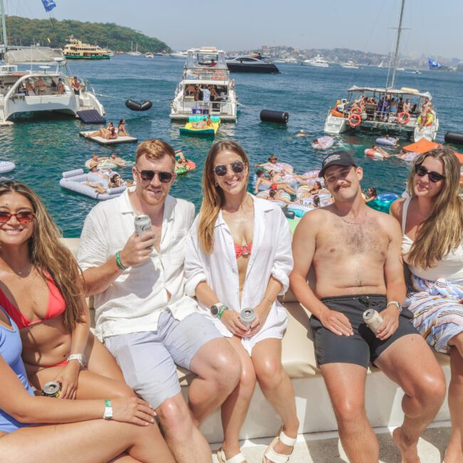 Six young adults wearing swimsuits and sunglasses sit on a white boat sofa, smiling at the camera. Behind them, people relax on floats and boats in a busy, sunny waterfront scene.